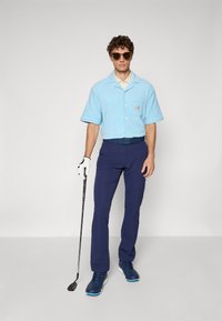 Férfi napszemüvegben és kék golf öltözetben, fehér kesztyűt viselve a bal kezén, egy golfütőt tartva, egyszínű háttér előtt áll.