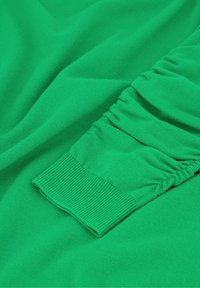 Groene gebreide stof met een geplooide mouwdetail en ribgebonden manchet. De textuur is zacht met een gladde afwerking.