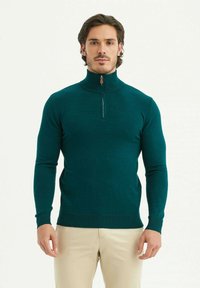 Pull en turquois en maille zippé, avec un col montant, des manches longues et un détail de fermeture éclair fine, porté avec un pantalon beige.