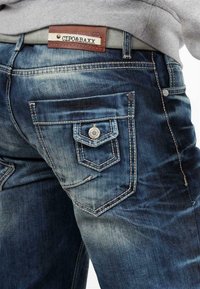 Jean en denim bleu avec une finition délavée, présentant des accessoires en métal argenté, des surpiqûres décoratives, et une étiquette en cuir marron au niveau de la taille.