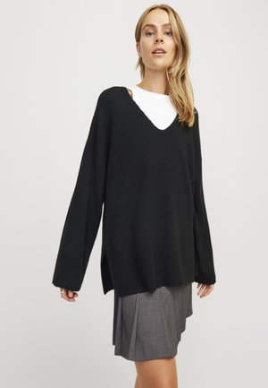 Svart oversized genser med V-hals og sideslit, laget av myk strikket materiale. Bæres over en hvit skjorte og kombinert med et grått skjørt.