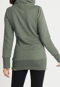 Sudadera con capucha de manga larga en verde oscuro, con dobladillo acanalado, que presenta un cuello enrollado y un pequeño parche de logo en la parte inferior.