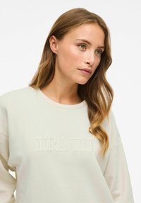 Felpa leggera beige con collo a giro, caratterizzata da testo in rilievo "TRUTH" sul davanti. Tessuto morbido e design dalla vestibilità rilassata.