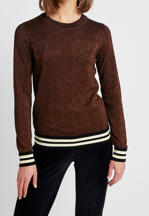 Brązowy sweter z długim rękawem z błyszczącej dzianiny z wzorem w zygzak oraz czarno-białymi paskowanymi mankietami i dolnym brzegiem, noszony z czarnymi prążkowymi spodniami.