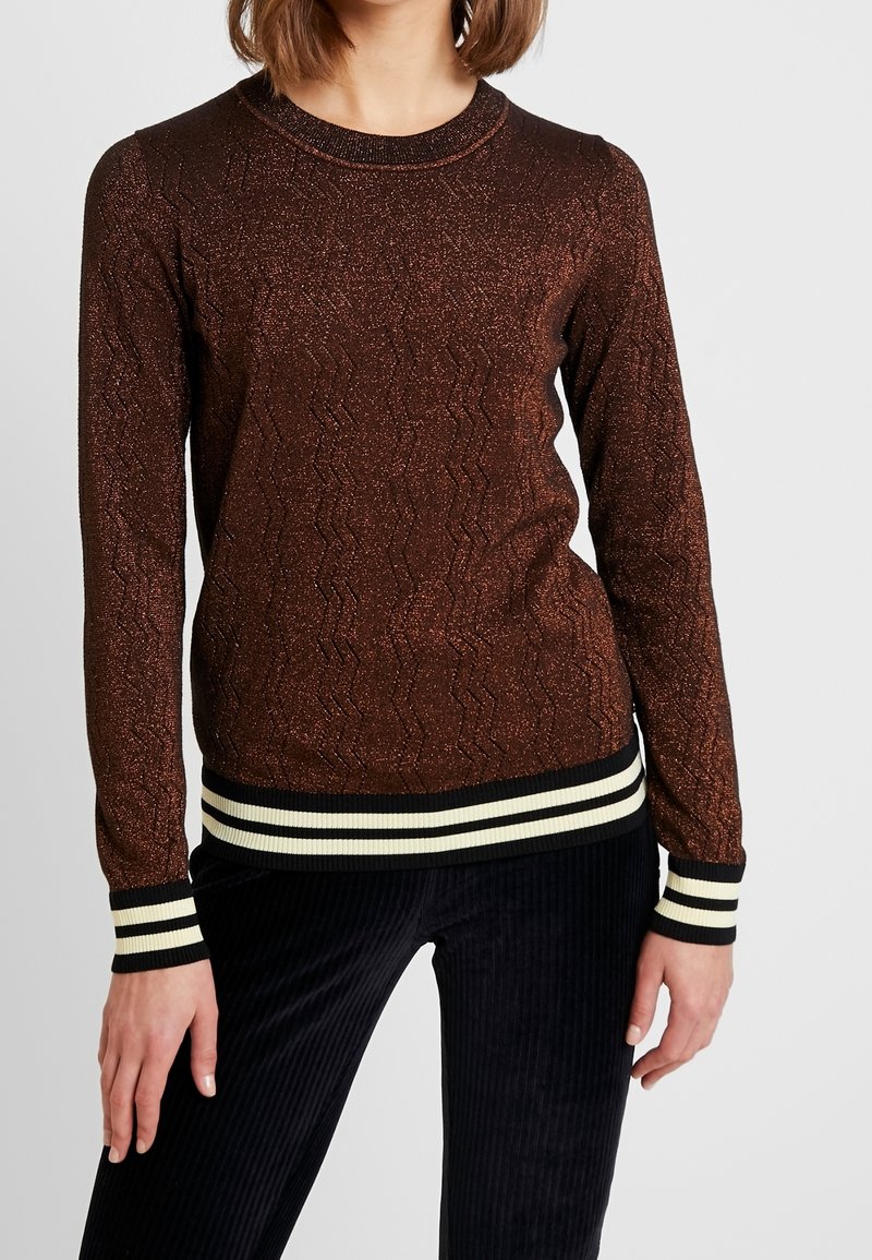 Pull en maille pailletée marron à manches longues avec motif en zigzag et poignets et ourlet rayés noirs et blancs, porté avec un pantalon noir côtelé.