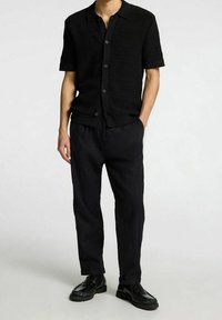 Camicia nera a maniche corte con trama, dotata di bottoni, abbinata a pantaloni neri larghi e scarpe nere stringate, creando un look monocromatico.