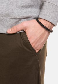 Sudadera gris con un acabado texturizado y un bolsillo; pantalones marrones con una superficie lisa. Una pulsera de cuerda negra en la muñeca.