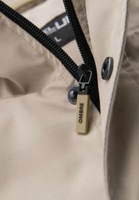 Beige Stoff mit einem teilweise geöffneten schwarzen Reißverschluss, versehen mit einem beigen Reißverschlusszug, der mit "OMBRE" beschriftet ist, sowie einem schwarzen Druckknopf.