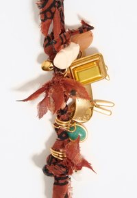 Charm de tono dorado sujetos a una cuerda trenzada de tela de color óxido. Incluye varias cuentas en crema, naranja, verde y un charm rectangular.