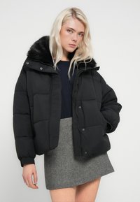 Crna jakna od puf materijala s umjetnim krznom na ovratniku, ima prošiveni dizajn, zatvarač i elastične manžete, uparena s sivom suknjom uzorka riblje kosti.