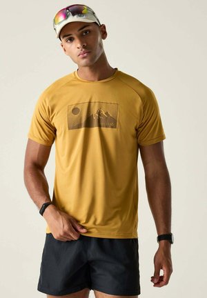 Junger Mann trägt ein senfgelbes T-Shirt mit Berg- und Sonnenmotiv, schwarze Shorts, eine weiße Kappe mit reflektierender Sonnenbrille und Fitnessbänder.