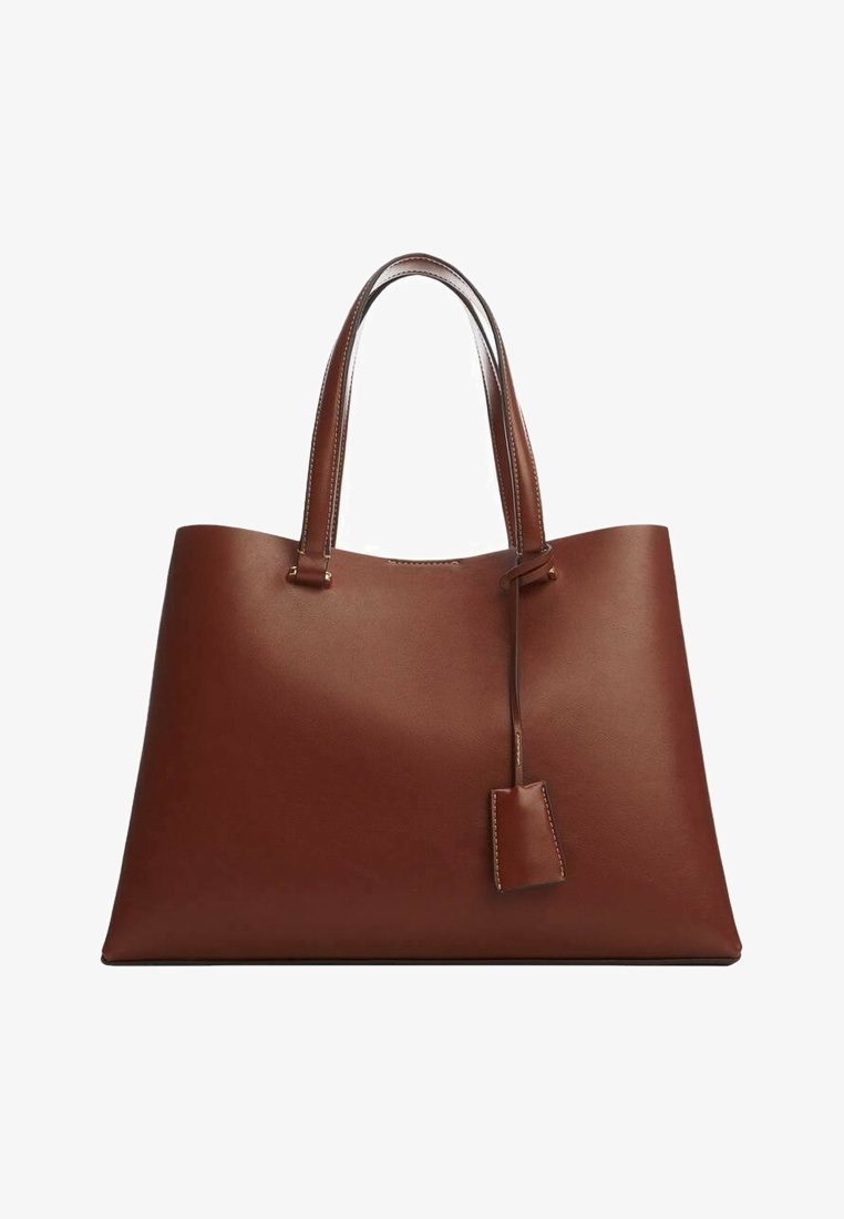 Grand sac fourre-tout en cuir marron avec deux poignées et une étiquette en cuir suspendue sur un fond blanc.