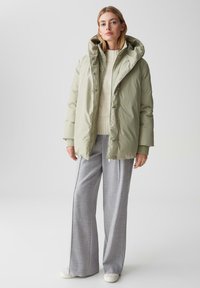 Giacca a vento verde chiaro con colletto alto e grande cappuccio, abbinata a un maglione crema a costine e pantaloni grigi a gamba larga.