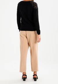 Suéter negro de manga larga con acentos de encaje en los hombros, combinado con pantalones de pierna ancha beige y sandalias negras de tacón. Se muestra vista trasera.