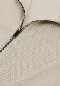 Beżowa prążkowana tkanina z częściowo odsłoniętym czarnym metalowym zamkiem błyskawicznym oraz małym wytłoczonym logo na powierzchni ubrania.