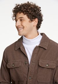 Camisa de pana marrón con dos bolsillos frontales y botones negros, superpuesta a un cuello de tortuga blanco. El cuello es acanalado y estructurado.