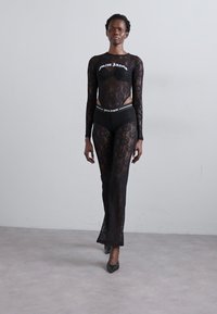 Black lace bodysuit with long sleeves and cutouts, paired with matching lace pants. Features a white logo and a fitted design.
