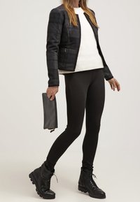 Giacca a scacchi nera e grigia, maglione bianco a maglia, leggins neri e stivali neri con lacci. Borsa a tracolla nera piegata tenuta in una mano.