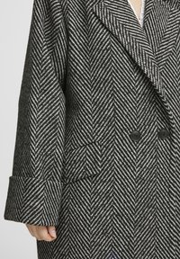 Cappotto doppiopetto in un motivo a spina di pesce bianco e nero, con ampie revers, due bottoni neri e maniche leggermente svasate.