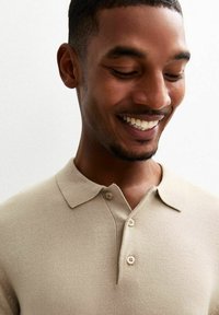 Beige Strick-Polohemd mit Kragen, kurzen Ärmeln, drei Knöpfen am Hals und einer weichen Textur. Das Modell lächelt, der Hintergrund ist schlicht.