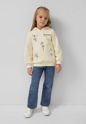 Crème hoodie met grafische afbeeldingen van een ballerina, borduurtekst "HET LEVEN IS BETTER als je danst," gecombineerd met blauwe flared jeans en witte sneakers.