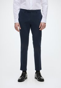 Sötétkék, szabott nadrág sima textúrával és slim fit fazonnal, fekete fűzős cipővel és fehér hosszú ujjú inggel párosítva.