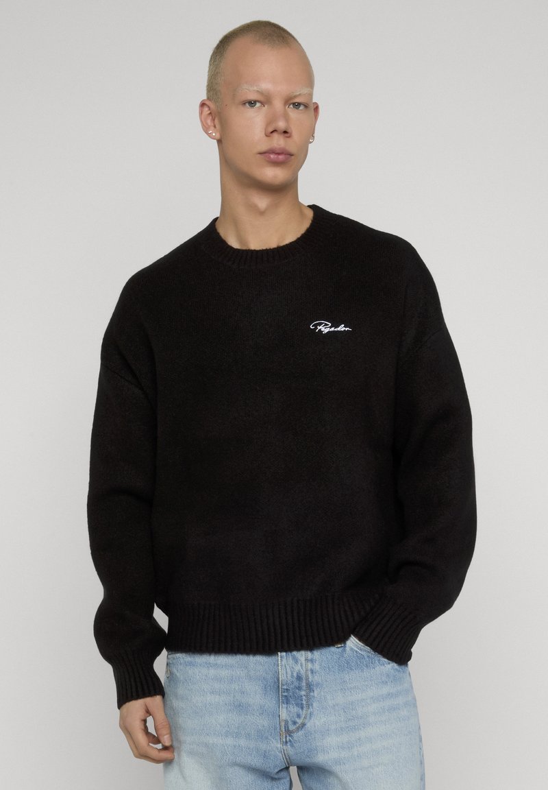 Jeune personne aux cheveux blonds courts portant un pull en maille noir avec un logo en script blanc et un jean bleu clair sur fond uni.