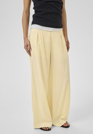 Személy széles szárú, világos sárga nadrágot visel fekete ujjatlan felsővel és fekete nyitott orrú szandállal, világosszürke padlón állva.