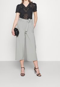 Kvinna i svart spetskortärmad topp, högmidjade ljusgrå vida cropped-byxor med bälte, svarta klackar med remmar, håller en svart clutchtvå.