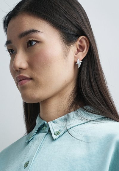 Stříbrné náušnice ve tvaru hvězdy s texturovaným povrchem, které má na sobě osoba v světle modré košili s límečkem. Viditelné jemné detaily vlasů.