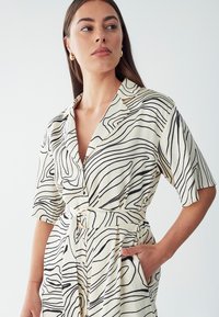 Korte mouw, beige jurk met zwarte, golvende lijnen. Heeft een ceintuur in de taille, een knopenlijst vooraan en zijzakken. Glad, lichtgewicht stof.