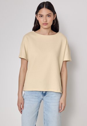 Haut beige à manches courtes côtelé avec un large col rond, associé à un jean en denim bleu clair. Le tissu a une finition douce et texturée.