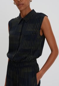 Sleeveless, collared shirt in navy with vertical olive green stripes, featuring a chest pocket and an elastic waistband for fit.