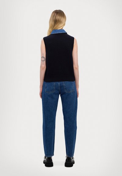 Mouwloze marineblauwe gebreide trui met een denim kraag, gecombineerd met hoog- taille lichtblauwe jeans en zwarte plateau-schoenen. Zichtbare tatoeage op de arm.