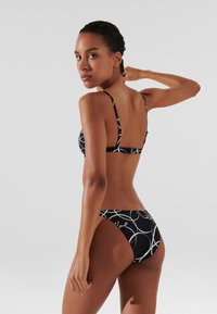 Čierny bikiny s bielym kruhovým vzorom, nastaviteľné tenké ramienka a zapínanie na chrbte. Materiál má hladkú textúru.