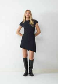 Czarna sukienka krótka z asymetrycznym dołem, wykonana z gładkiego materiału. Nosi ją z wysokimi, czarnymi botkami na tle neutralnym.