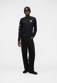 Maglietta nera a maniche lunghe con grafiche arancioni sulle maniche; abbinata a pantaloni neri e un berretto scuro; modello in piedi con le mani nelle tasche.