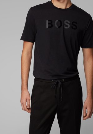 Homme portant un T-shirt noir à manches courtes avec l'inscription brillante "BOSS" et un pantalon noir à cordon de serrage, sur un fond gris uni.