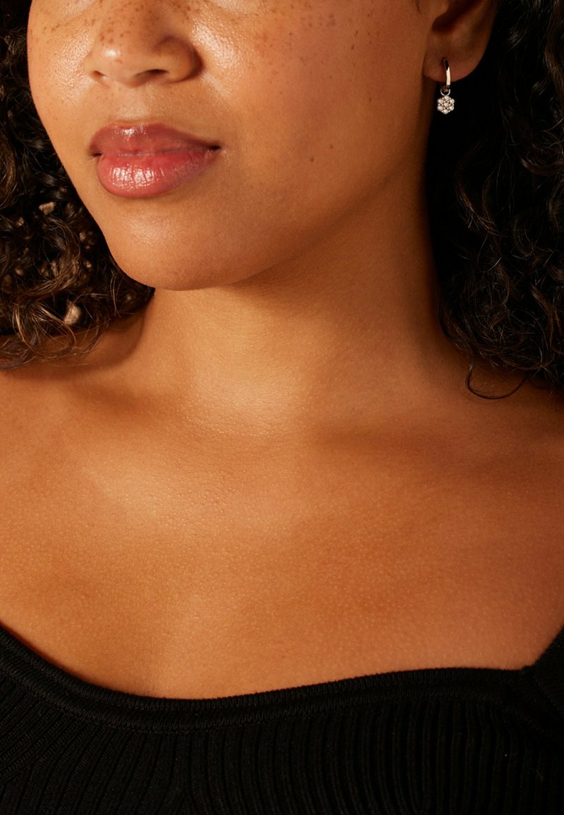 Boucle d'oreille en argent en forme d'anneau avec un motif floral, portée par une personne aux cheveux bouclés. Haut noir à texture côtelée. Peau lisse visible.