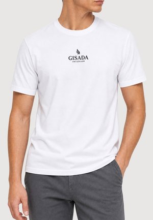 Mann trägt ein weißes T-Shirt mit kurzen Ärmeln und "GISADA Switzerland"-Logo auf der Brust und dunkelgraue Hose, eine Hand in der Tasche.