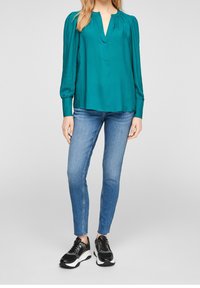 Blusa teal con maniche lunghe e gonfie e scollatura a V, abbinata a jeans blu aderenti e sneakers nere. Tessuto morbido, vestibilità comoda.