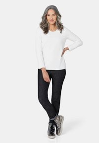 Weißes Langarmoberteil, dunkle Jeans und metallische High-Top-Sneaker. Das Model steht mit einer Hand auf der Hüfte und zeigt eine lockere Passform.