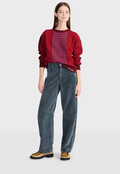 Jeune femme portant un pull rouge à motifs, un pantalon ample en velours côtelé gris et des chaussures bicolores beige et noir, debout devant un fond uni.