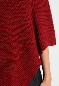 Pull rouge en tricot avec texture côtelée diagonale porté sur le bras, associé à un pantalon noir avec une poche zippée visible.