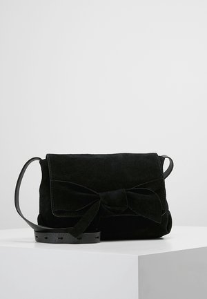 Schwarze Wildleder-Schultertasche mit Frontklappe und großer dekorativer Schleife, die auf einem weißen Podest vor neutralem Hintergrund ruht.