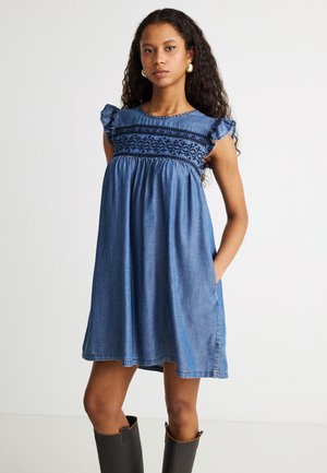 Joven con cabello largo y rizado que lleva un vestido de mezclilla azul bordado con mangas con volantes y botas negras hasta la rodilla.