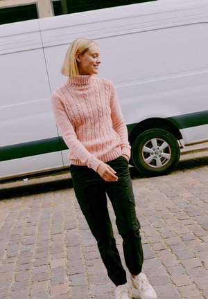 Rosa kabelstickad polotröja, svarta cargobyxor och vita sneakers. Modellen står på kullerstenar med en vit skåpbil i bakgrunden.