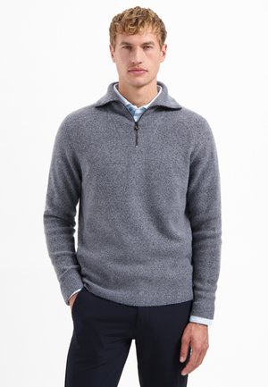 Man in een lichtblauw overhemd met kraag onder een grijze kwart-rits sweater en donkere broek, staande met één hand in zijn zak.