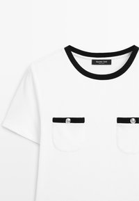 White short-sleeve shirt with black trim on neckline and pocket tops, featuring silver buttons on both chest pockets.