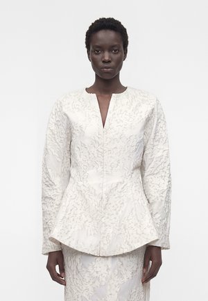 Kobieta o krótkich kręconych włosach, ubrana w biały teksturowany blezer z długim rękawem i pasującą spódnicę, stojąca na tle jednolitego białego tła.